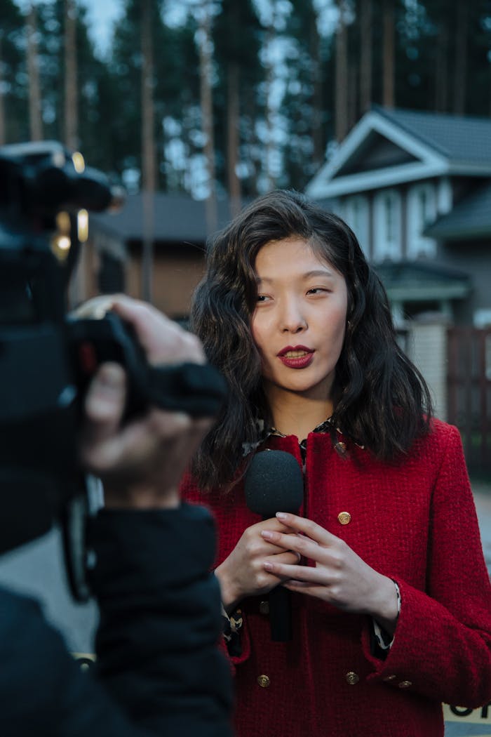 Asian woman reporting news on camera outdoors, wearing red coat.