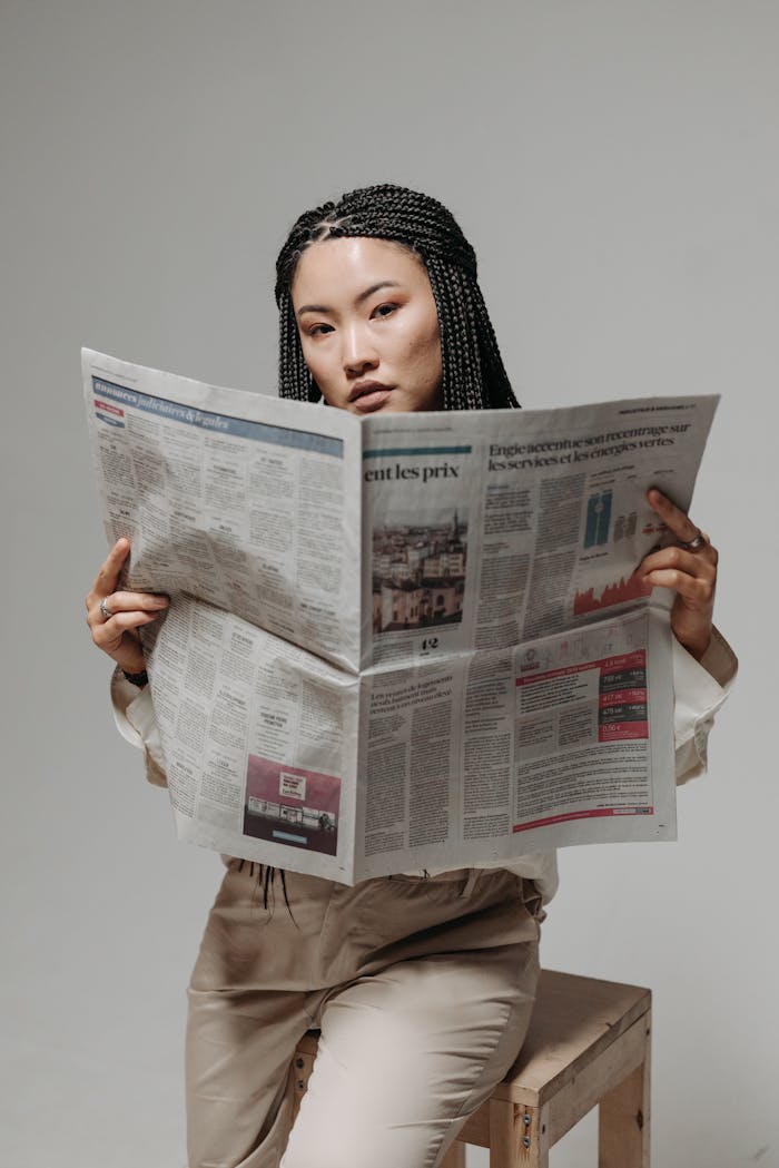 A stylish woman with braided hair reading a newspaper in a studio setting.